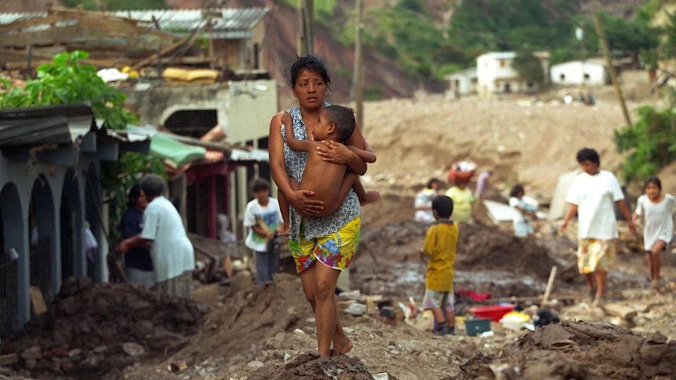 La imagen muestra a supervivientes del huracán Mitch en medio de la destrucción. En el medio, una mujer carga a un bebé desnudo mientras camina por los escombros.
