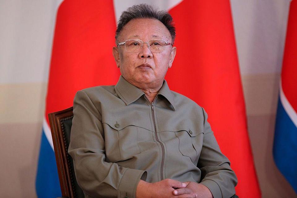 El exlíder de Corea del Norte, Kim Jong Il, está sentado en una silla con una camisa verde y gafas, con banderas de Corea del Norte en el fondo.