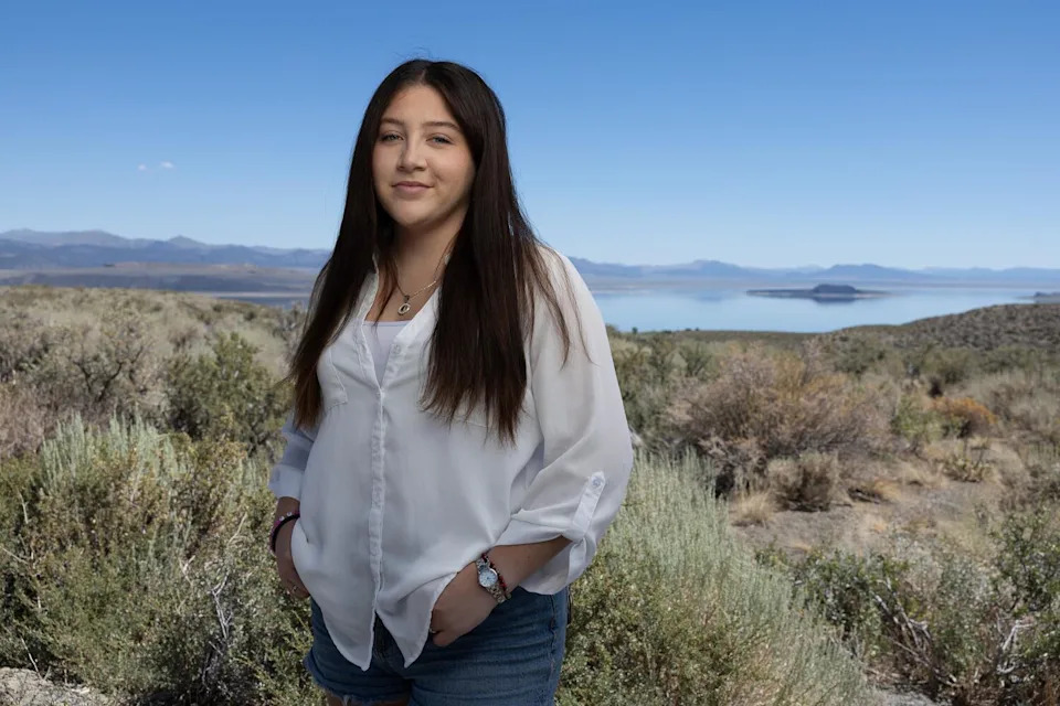 A young woman with long dark hair, in a white long-sleeved shirt and jeans, standing amid shrubs near a lake
