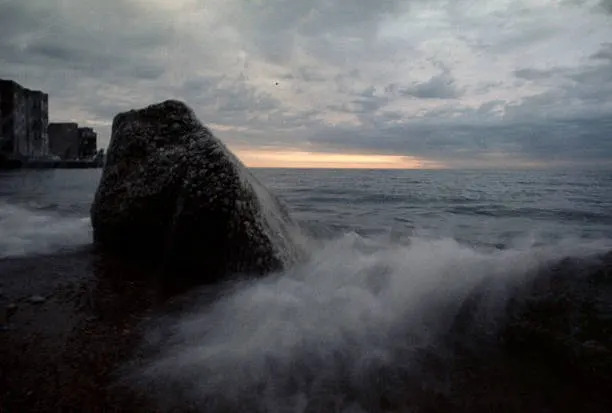 Waves crash against a large rock on a pebble beach, with a cloudy sky and distant horizon in the background
