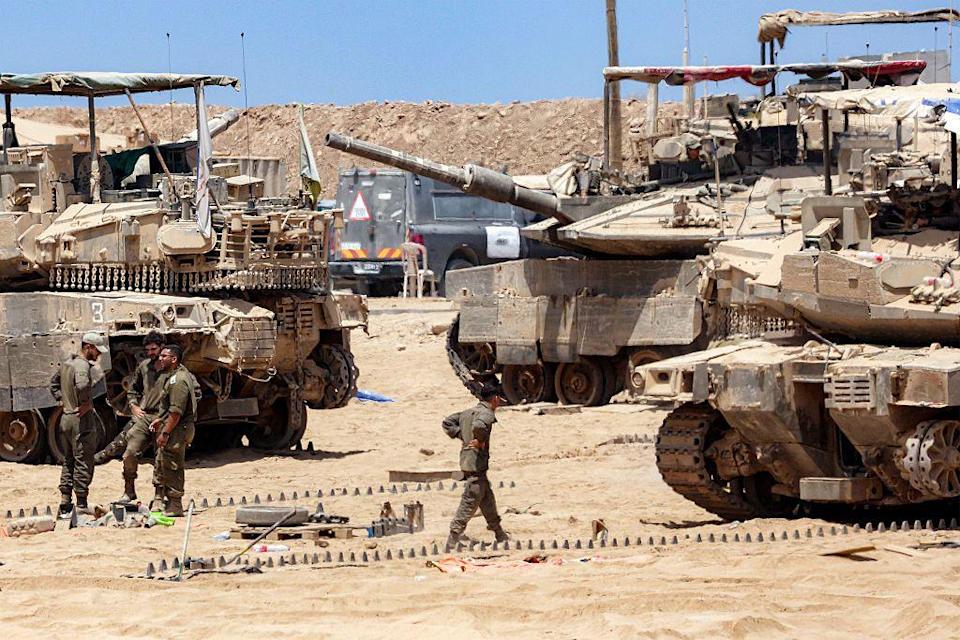 Un soldado israelí camina entre tres tanques de guerra en el desierto, mientras otros tres soldados están parados en un grupo hablando y riendo.