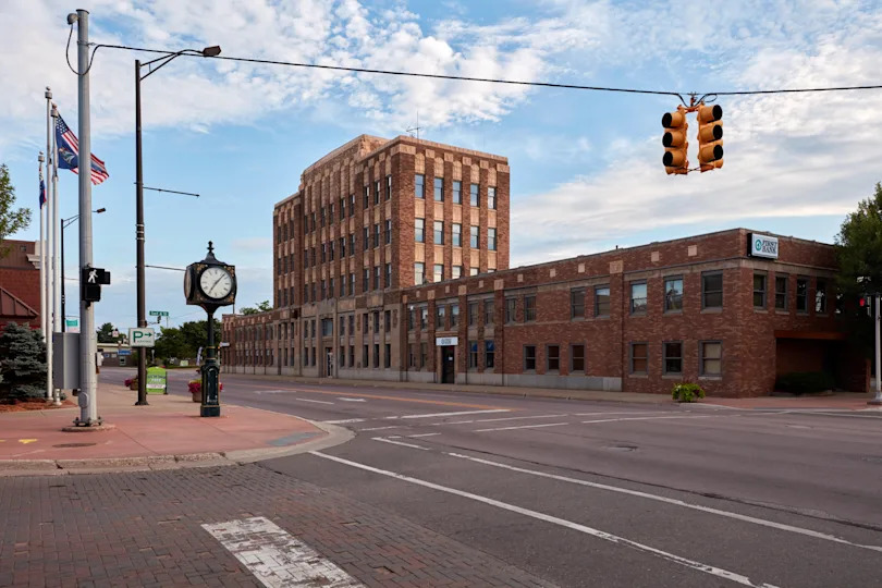 A brick building on a quiet street corner with a clock and traffic light. Flags and a tree are visible, with a cloudy sky above