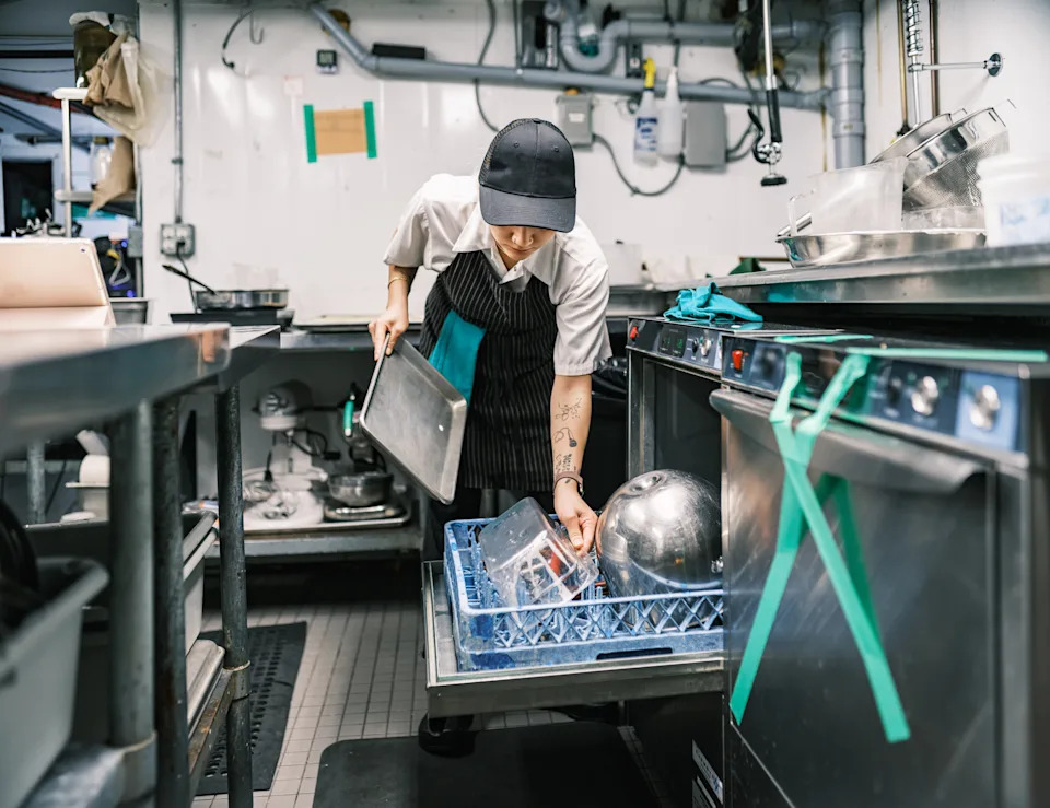 Person in a commercial kitchen loading dishes into an industrial dishwasher. Wearing a cap, striped apron, and white shirt. Working environment