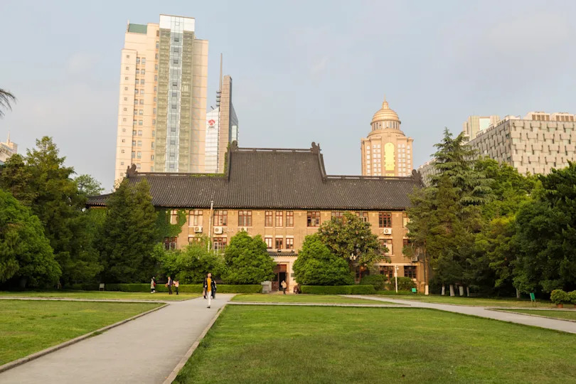 Historic building with a traditional roof, surrounded by grass and trees, stands among modern skyscrapers in the background