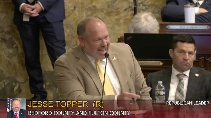 Jesse Topper speaks at a podium in a suit. A nameplate identifies him as representing Bedford and Fulton County. Audience members listen attentively