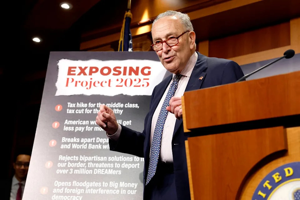 Senate Majority Leader Chuck Schumer stands near a sign titled "Exposing Project 2025" detailing various criticisms related to tax and border policies