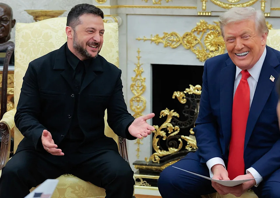 Two men in a formal setting, smiling and sitting on yellow armchairs, dressed in a black jacket and a blue suit with a red tie, respectively