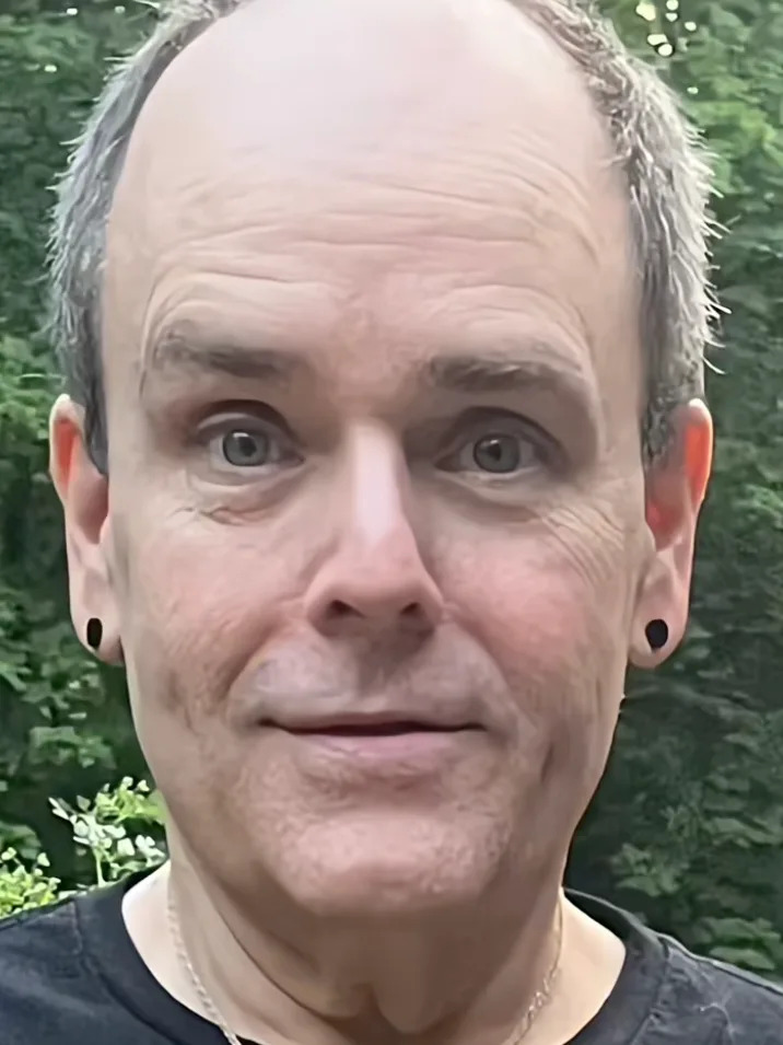 Man with short hair, wearing earrings and a black shirt, standing outdoors with greenery in the background