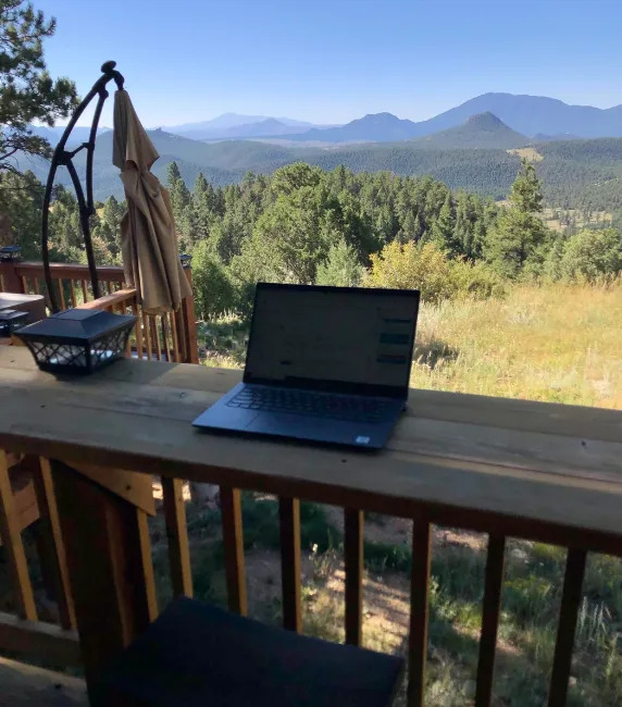 Laptop on a wooden deck overlooks a serene view of forested mountains under a clear sky. An umbrella and lanterns are nearby