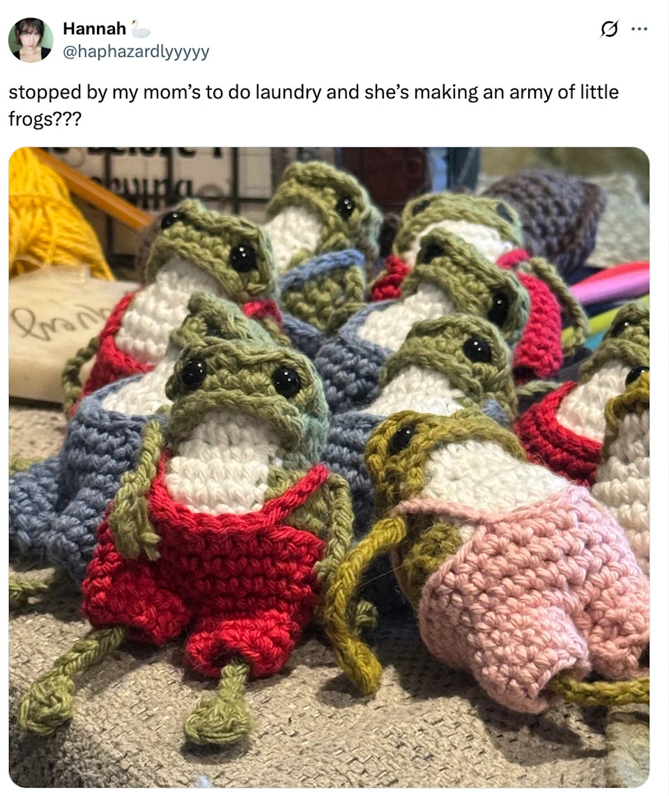 Crocheted frogs wearing shorts are displayed on a table, surrounded by yarn and crafting materials
