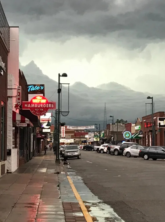Cloudy sky above a quiet street lined with parked cars and storefronts, including a hamburger restaurant, suggesting an impending storm
