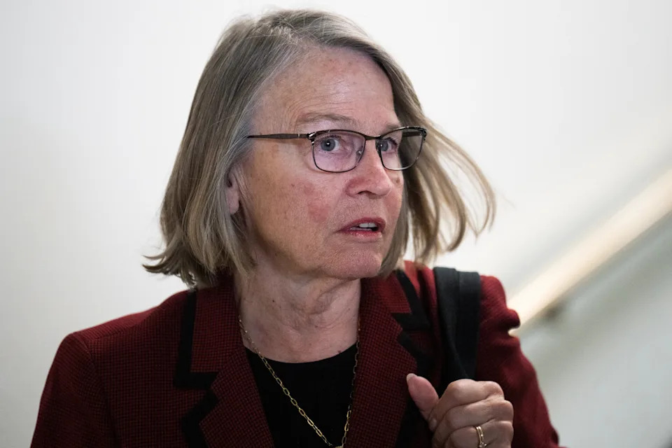 A person with medium-length hair wears glasses and a professional outfit, carrying a bag. They appear to be in an indoor setting