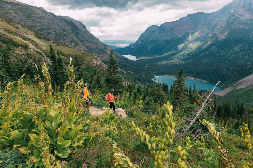 Two hikers on a trail in a mountainous landscape with lush greenery and lakes in the valley below