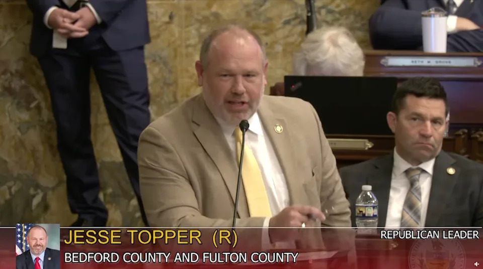 Jesse Topper speaks at a podium in a legislative setting; name and title displayed on the screen below him