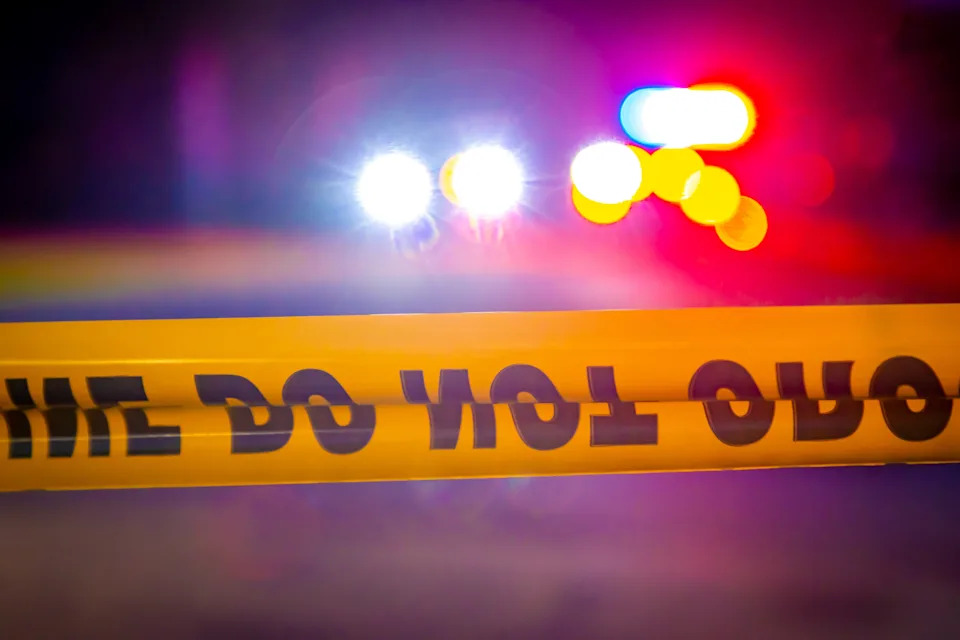 Blurred police lights behind yellow "Do Not Cross" tape, suggesting a crime scene at night