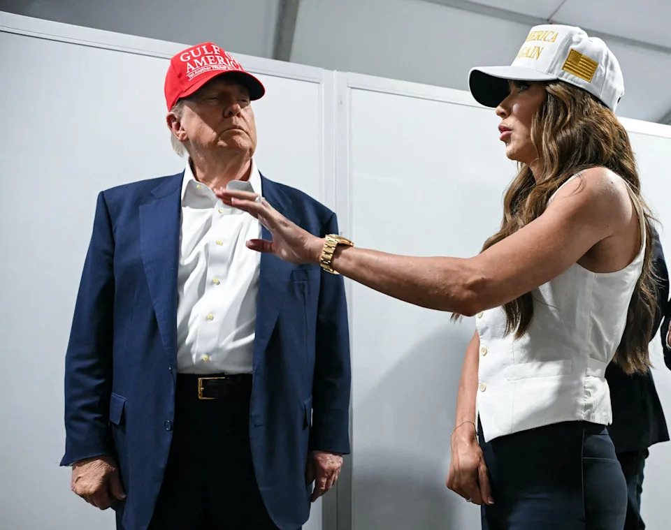 Two individuals are conversing. One wears a suit and a hat, while the other wears a sleeveless top and hat. A third person in a suit observes