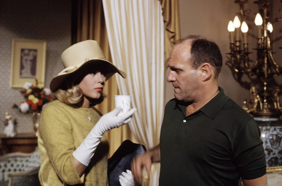 Woman in vintage style clothing and hat holds a cup, talking to a man in a casual shirt in an elegant room