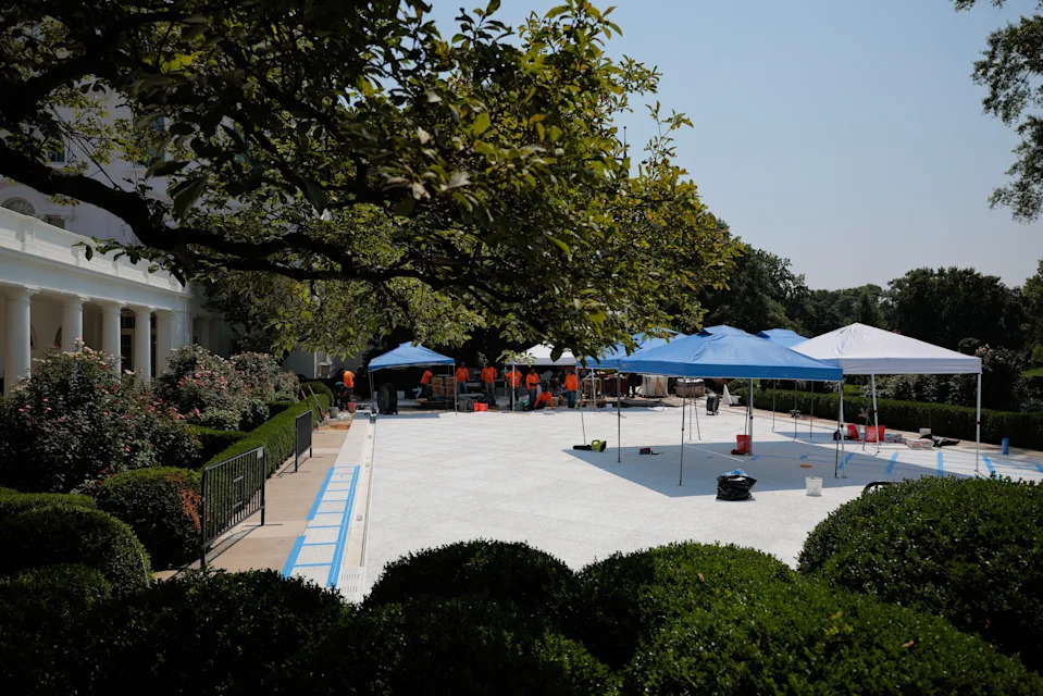 View of a construction project with white tents and workers on a spacious lawn, surrounded by lush trees and greenery