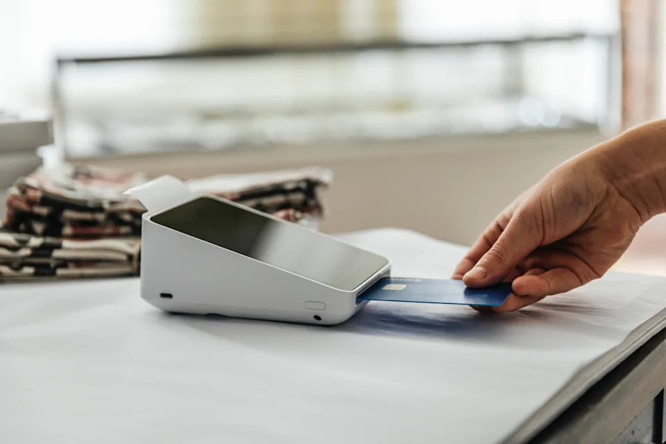 Hand inserting a credit card into a card reader on a counter with folded newspapers in the background, illustrating a transaction process