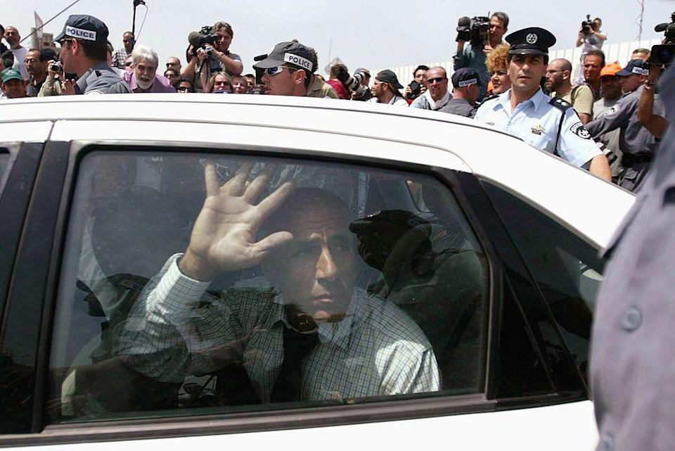 El denunciante israelí Mordechai Vanunu sale de prisión en un coche blanco, vestido con una camisa a cuadros y corbata negra, con la mano en la ventanilla del coche, mientras una multitud de periodistas y policías rodean su coche.