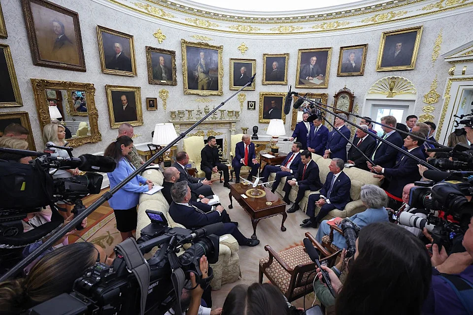 Media and officials gather in a decorated office for a meeting. Several cameras and microphones capture the scene. The room has many framed portraits