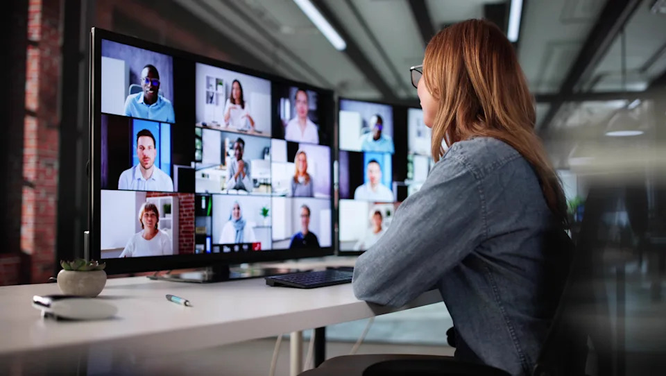 Person at desk in a virtual meeting with large monitor showing multiple participants, highlighting remote work collaboration