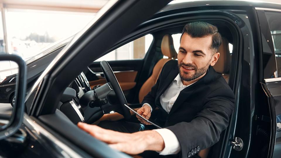A salesman in a suit with a tablet standing in a car showroom and demonstrating a car to buyers. Buying, renting and insuring a car.