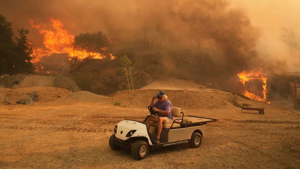 A man drives in a golf cart as a wildfire burns behind him