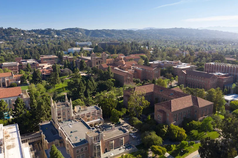 Aerial view of a university campus with historic brick buildings, surrounded by trees and set against a backdrop of mountains