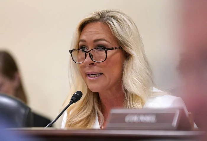 A person wearing glasses speaks into a microphone during a formal meeting or hearing, with blurred individuals in the background