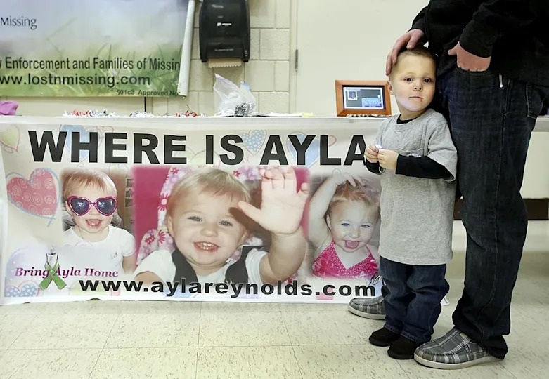 A child stands near an adult, holding a toy; a banner behind them displays photos of a missing girl with the text "WHERE IS AYLA?" and website details
