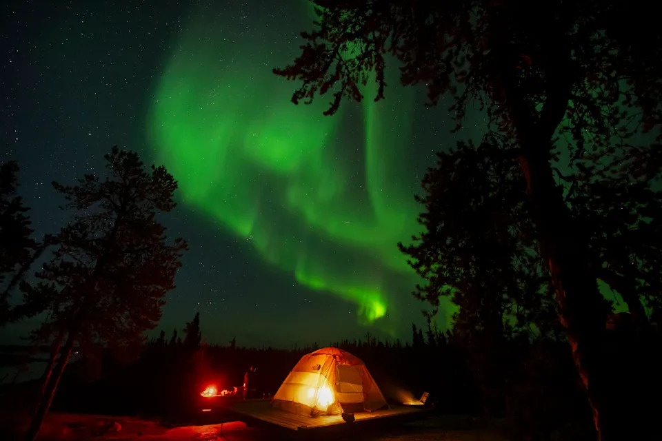 The northern lights glow green in the sky above an illuminated tent.