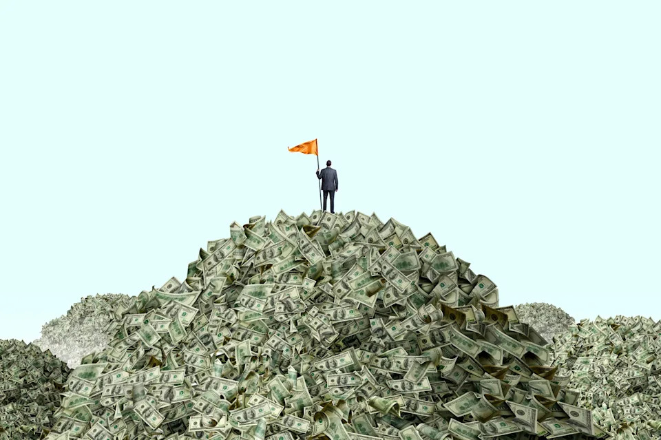 Person in a suit stands atop a large pile of money holding a flag, symbolizing financial success or conquest
