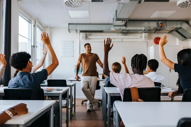 Students in a classroom raising hands, engaged with a teacher standing at the front, leading a discussion