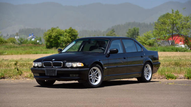 a black 2001 BMW 750iL parked in a parking lot on a smoggy day, with mountains and a red building in the background
