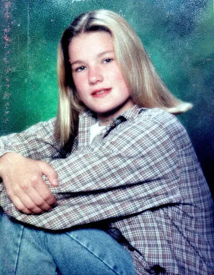 A person with shoulder-length hair sits casually with a relaxed expression, wearing a plaid shirt and jeans