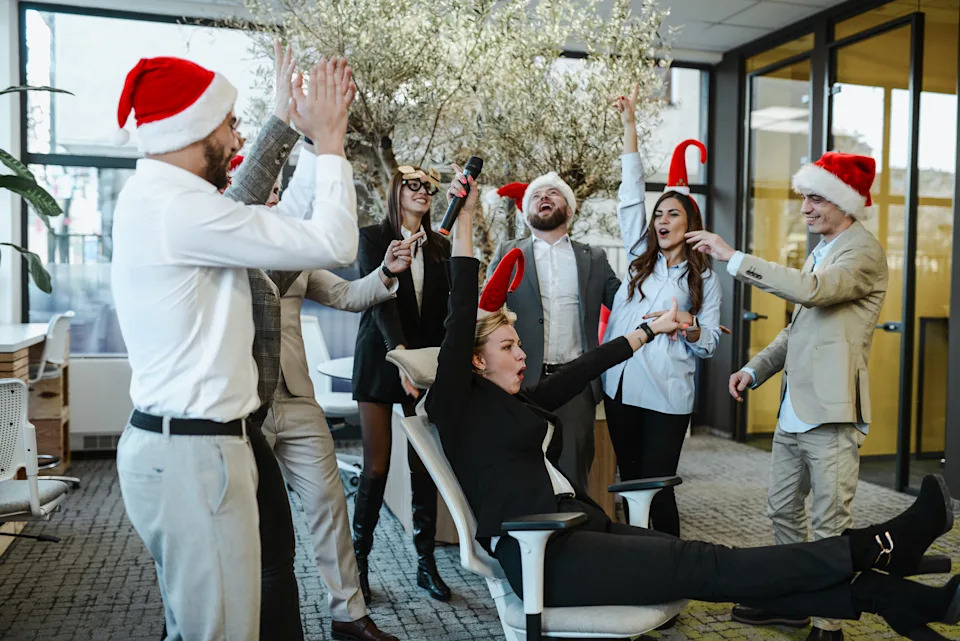 Office holiday party with people in Santa hats. One person joyfully spins in a chair while others cheer and celebrate around them