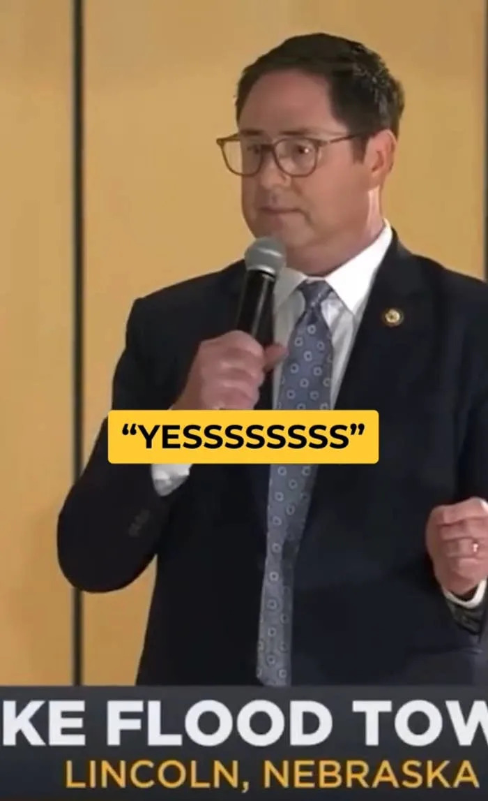 Man in a suit and tie holds a microphone, text reads "YESSSSSSS" and mentions "Lake Flood Town Hall, Lincoln, Nebraska" at the bottom