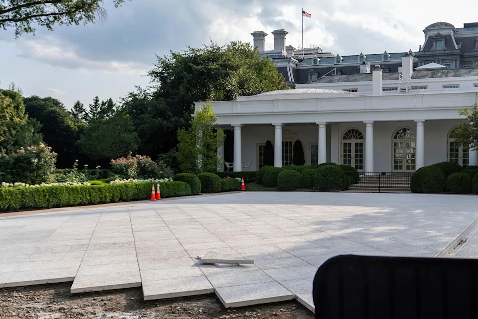 White House with partially constructed patio, surrounded by greenery, under a cloudy sky. Orange cones mark off a section of the area