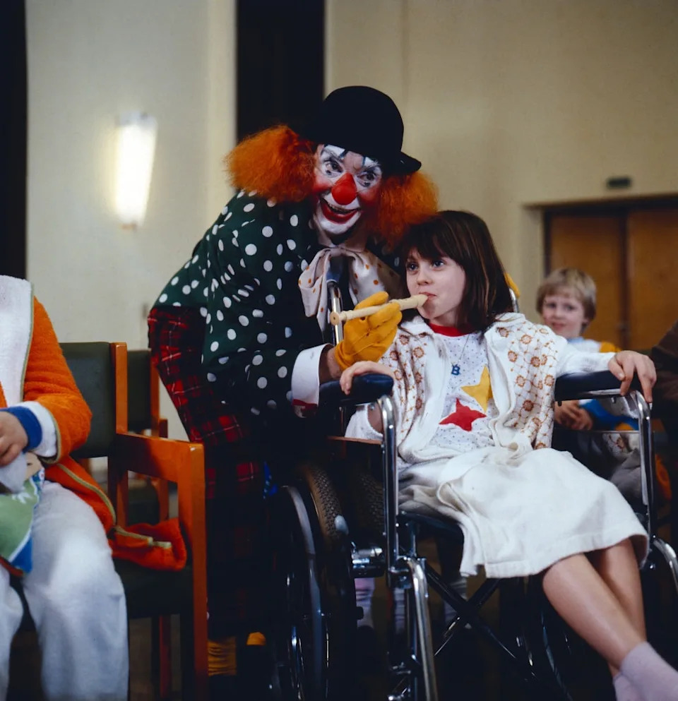 A clown entertaining a young girl sitting in a wheelchair at an indoor event, bringing joy and laughter to a group of children around them