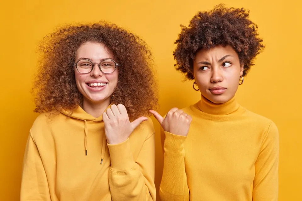 Two girlfriends pointing to one another.