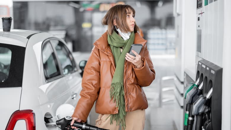 A woman in a brown coat and green scarf pumps gas into a white car in winter while holding her phone