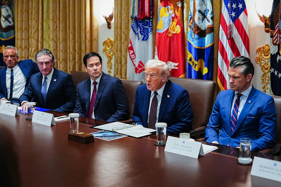A group of men seated at a long table in a formal meeting room with flags in the background