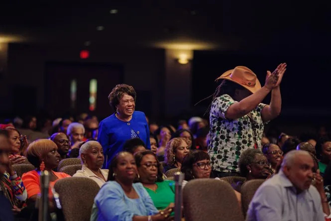 Two people in a crowd of seated people stand up smiling and clapping