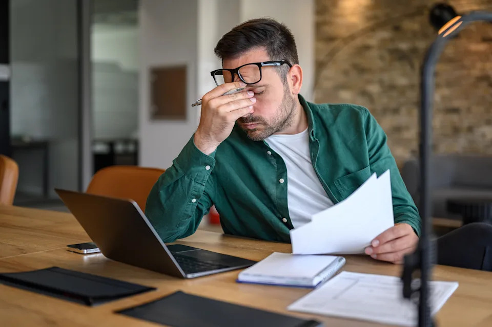 Person in an office rubbing eyes while holding papers, sitting at a desk with a laptop and open notebook, appearing tired or stressed