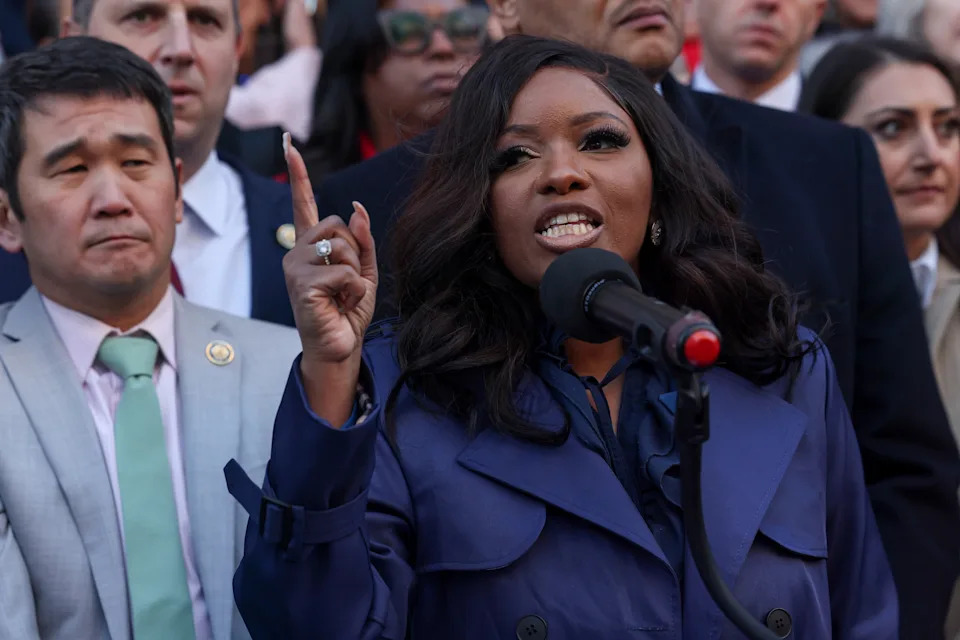 Person speaking passionately at a microphone at an outdoor event, surrounded by individuals in professional attire