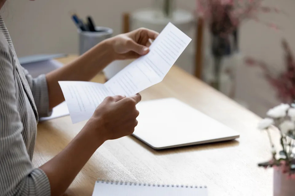 Person seated at desk, holding and reading a sheet of paper. Nearby notebook and pens visible, with soft focus on background decor