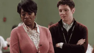 Two people in an indoor setting; one with a thoughtful expression, arms crossed, and the other with a skeptical look, shrugging slightly