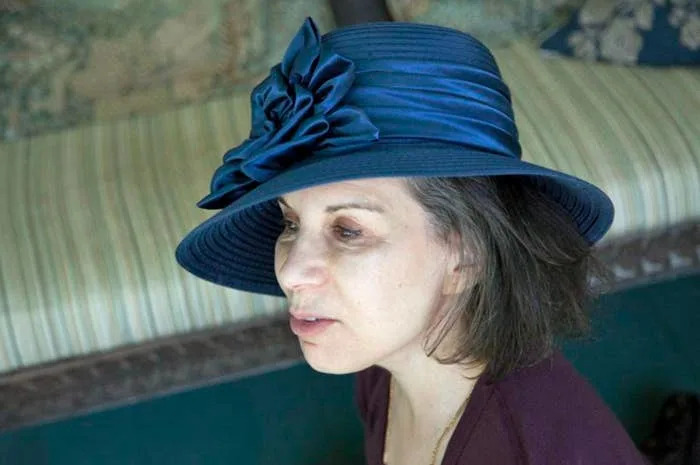 Person wearing a stylish wide-brimmed hat with a large ribbon accent, sitting indoors, looking thoughtful