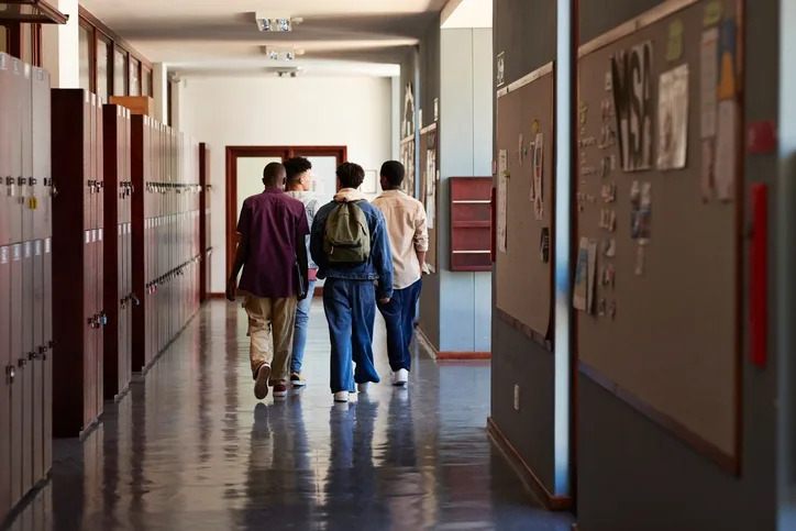 Students walking down a school hallway with lockers on the left and bulletin boards on the right. They appear to be engaged in conversation
